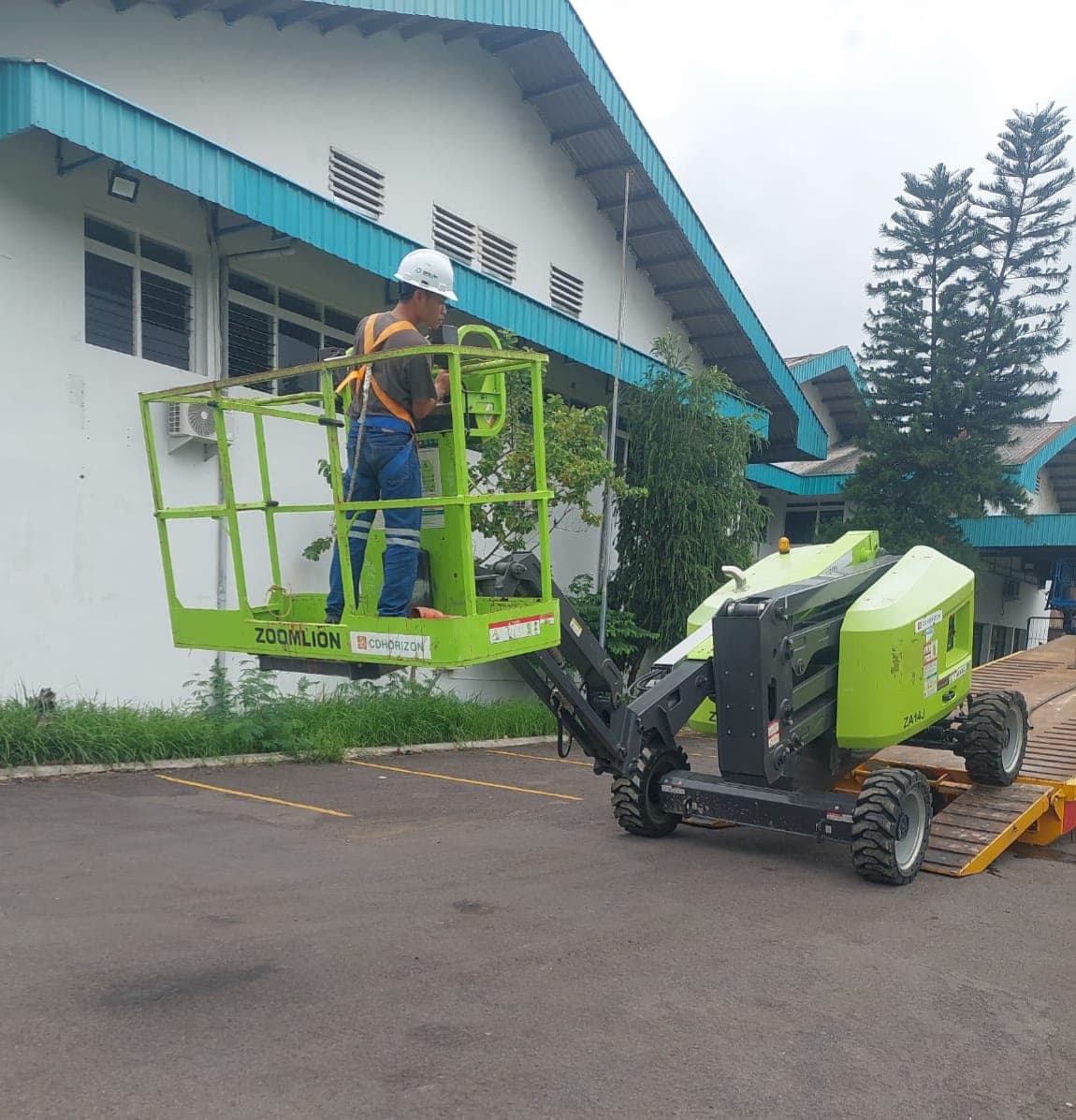 Pekerja dengan alat pengaman berdiri di keranjang boom lift Zoomlion hijau di atas ramp.