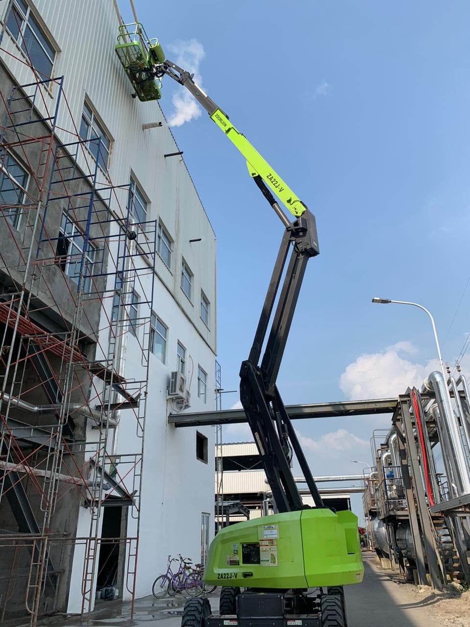 Boom lift hijau menjulang tinggi ke arah dinding bangunan industri putih di bawah langit biru.