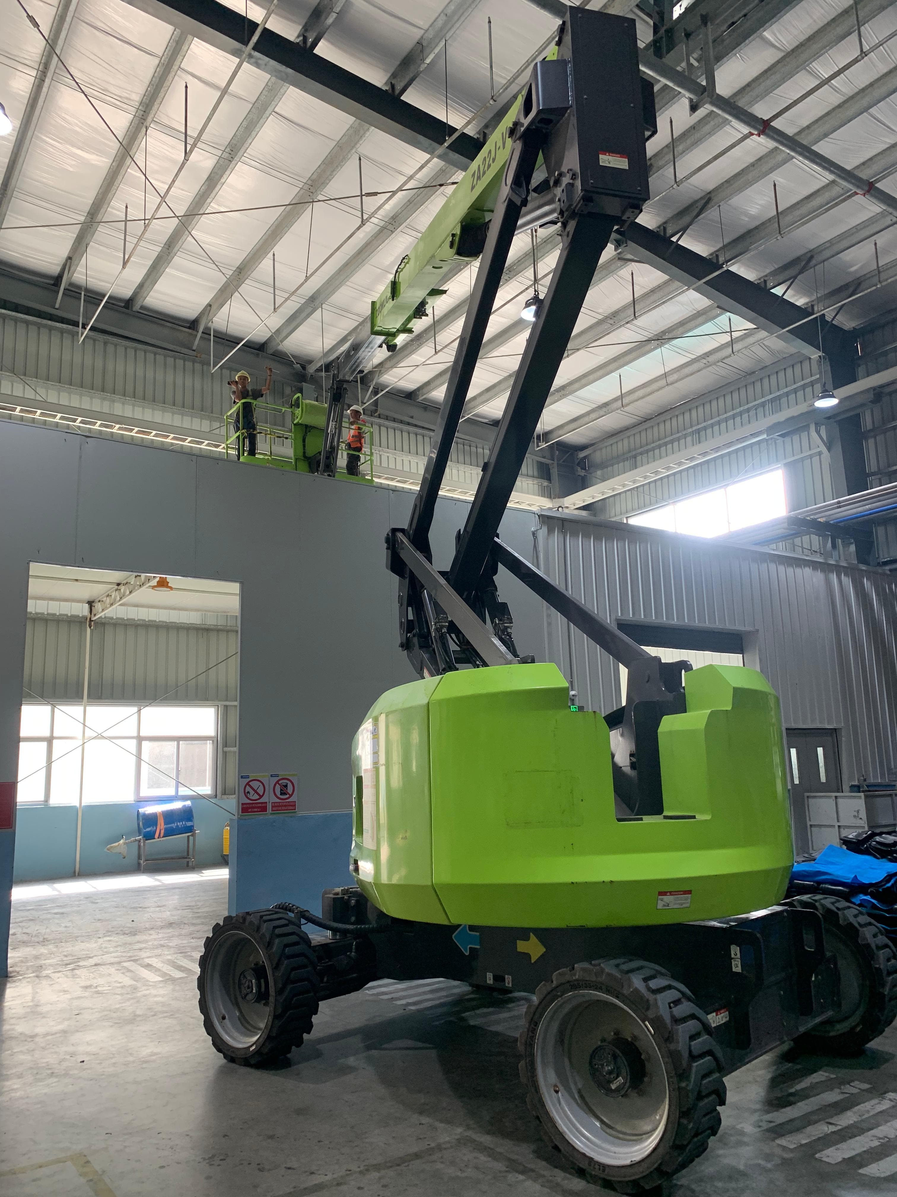 Boom lift hijau mengangkat dua pekerja ke langit-langit tinggi di dalam sebuah gudang industri.