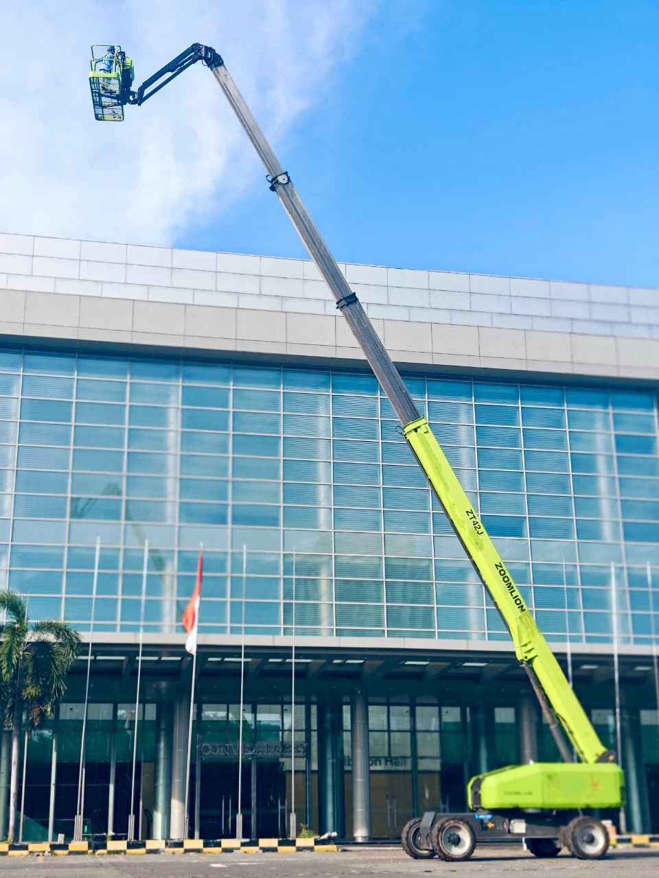 Boom lift Zoomlion hijau menjulang tinggi di depan gedung kaca dengan pekerja di dalam keranjang.