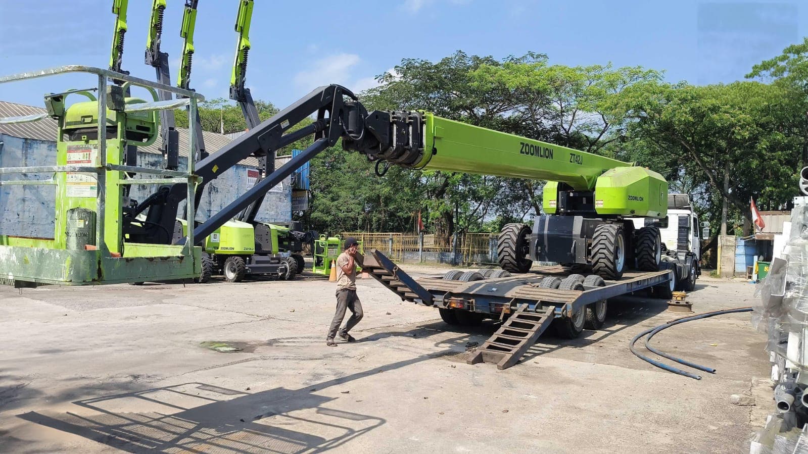 Boom lift Zoomlion hijau sedang dimuat ke atas truk trailer di lapangan terbuka saat siang.